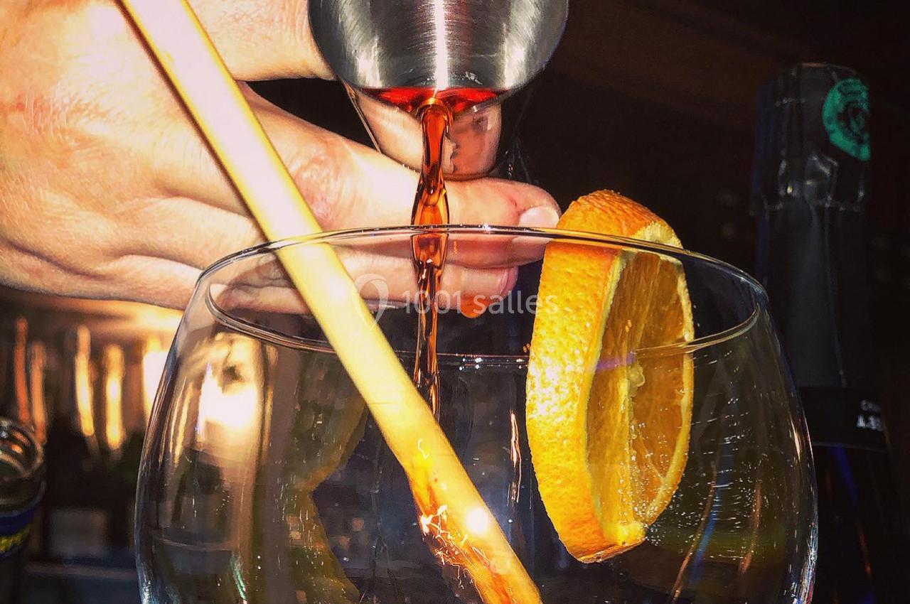 Main versant un liquide ambré dans un verre contenant une tranche d'orange et une décoration en bois.