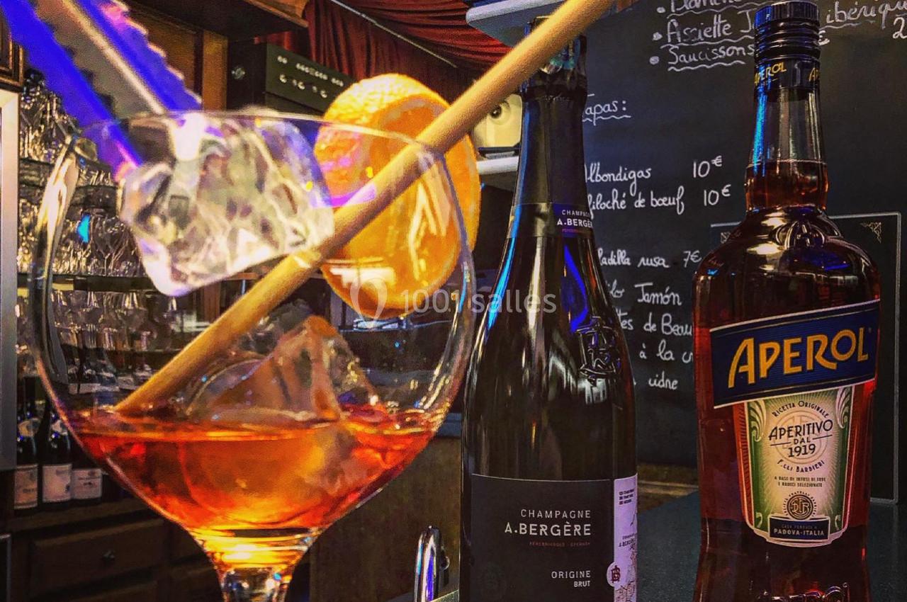 Verre de cocktail avec tranche d'orange et glaçons, accompagné de bouteilles de champagne et d'Aperol sur un bar.