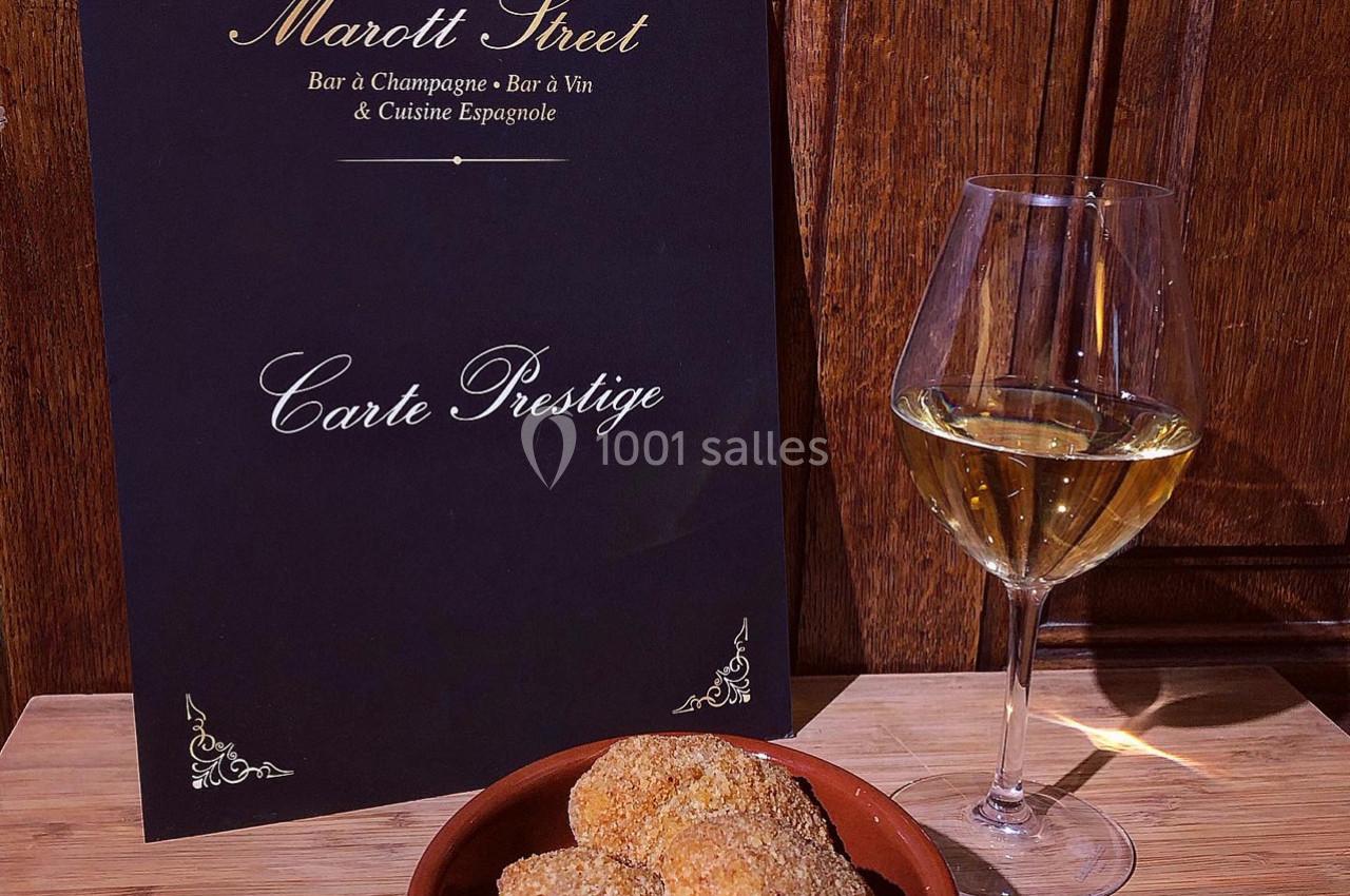 Carte de restaurant posée sur une table en bois avec un verre de vin blanc et une assiette de croquettes.