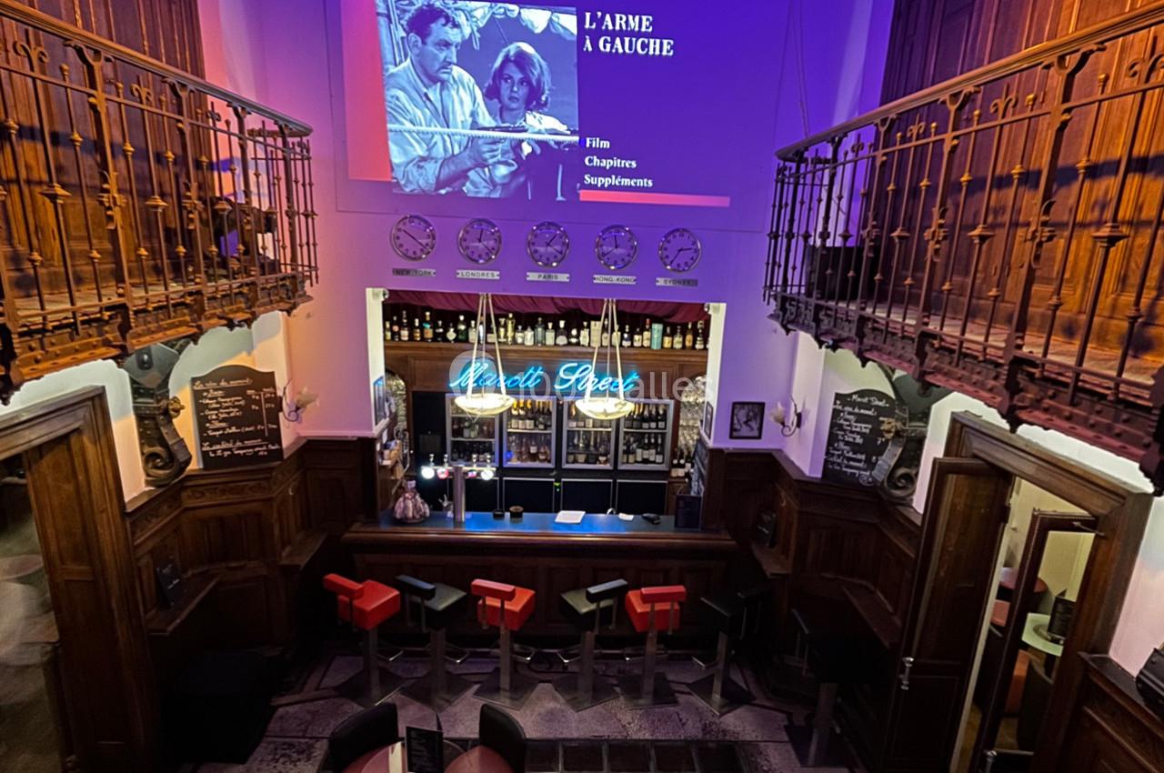 Vue intérieure d'un bar avec un comptoir éclairé, des tabourets rouges et un écran projetant un film en arrière-plan.