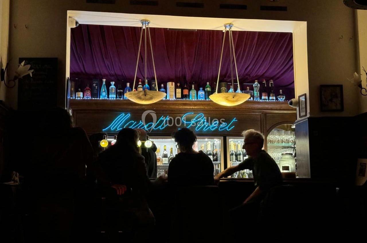 Un bar avec des étagères éclairées, des bouteilles alignées et des clients assis au comptoir dans une ambiance tamisée.