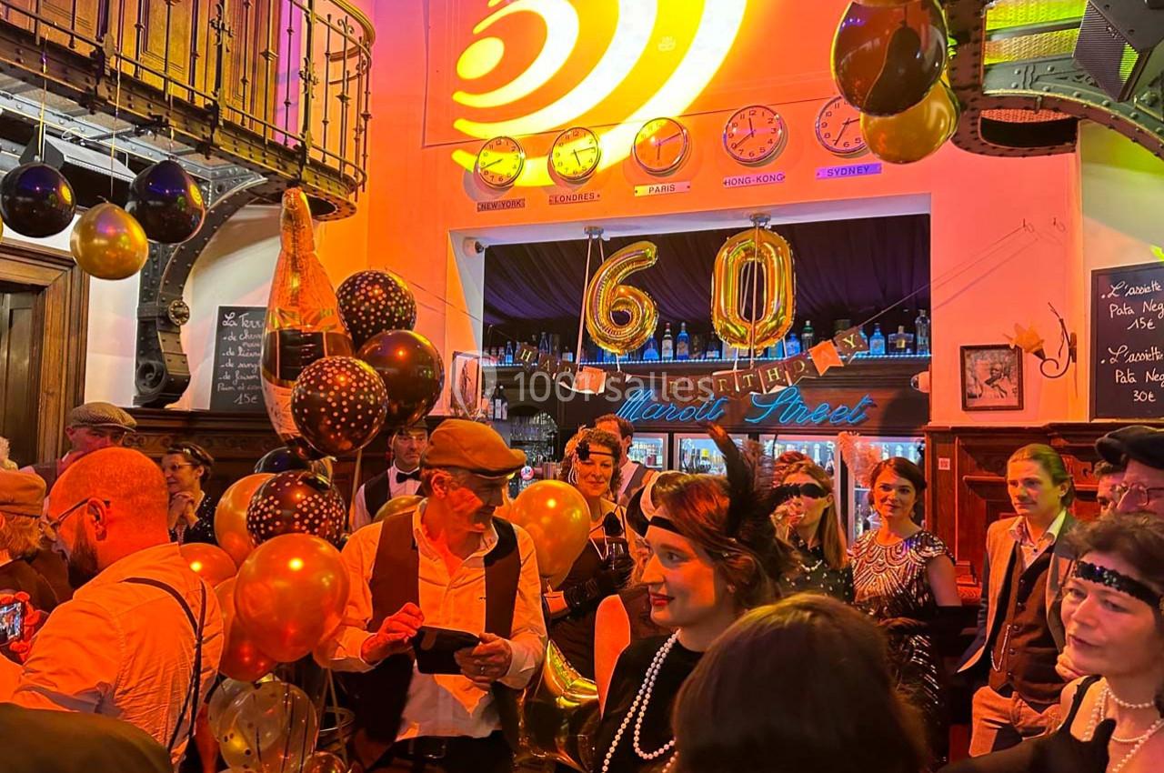 Personnes en tenue rétro célébrant un 60e anniversaire dans un bar décoré de ballons et lumières chaleureuses.