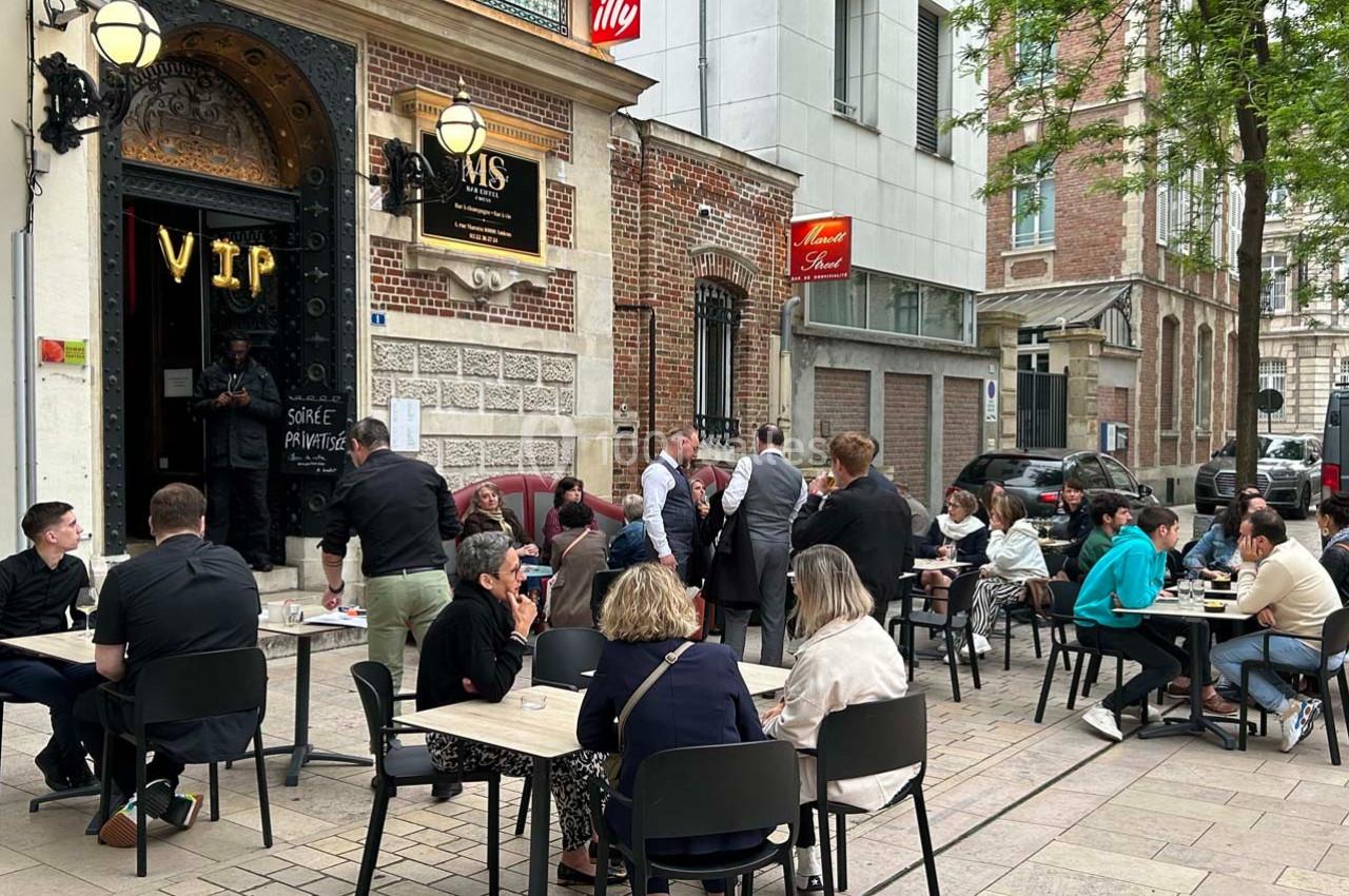 Des personnes assises en terrasse et debout devant un café-restaurant dans une rue urbaine animée.