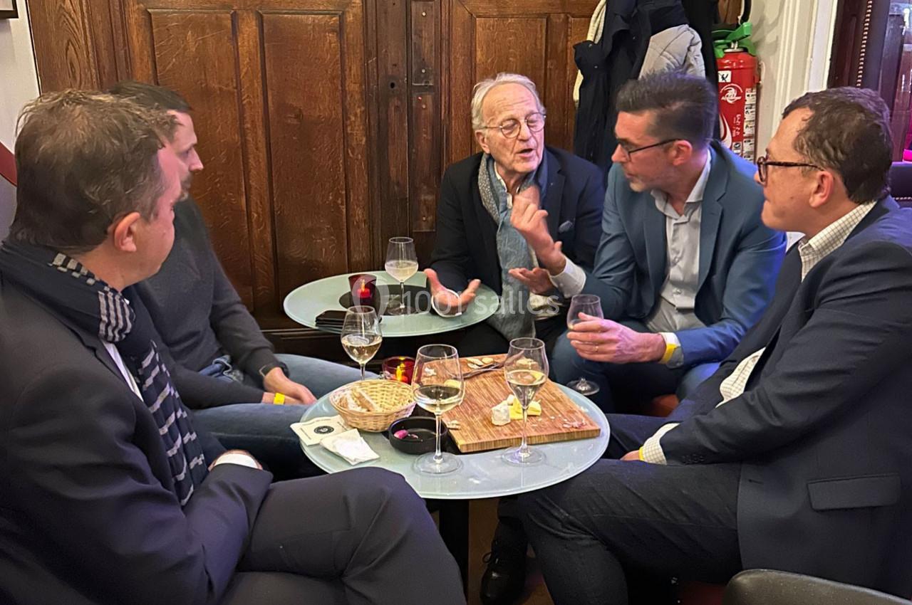 Cinq hommes discutent autour de petites tables avec des verres, des snacks et un décor intérieur en bois.