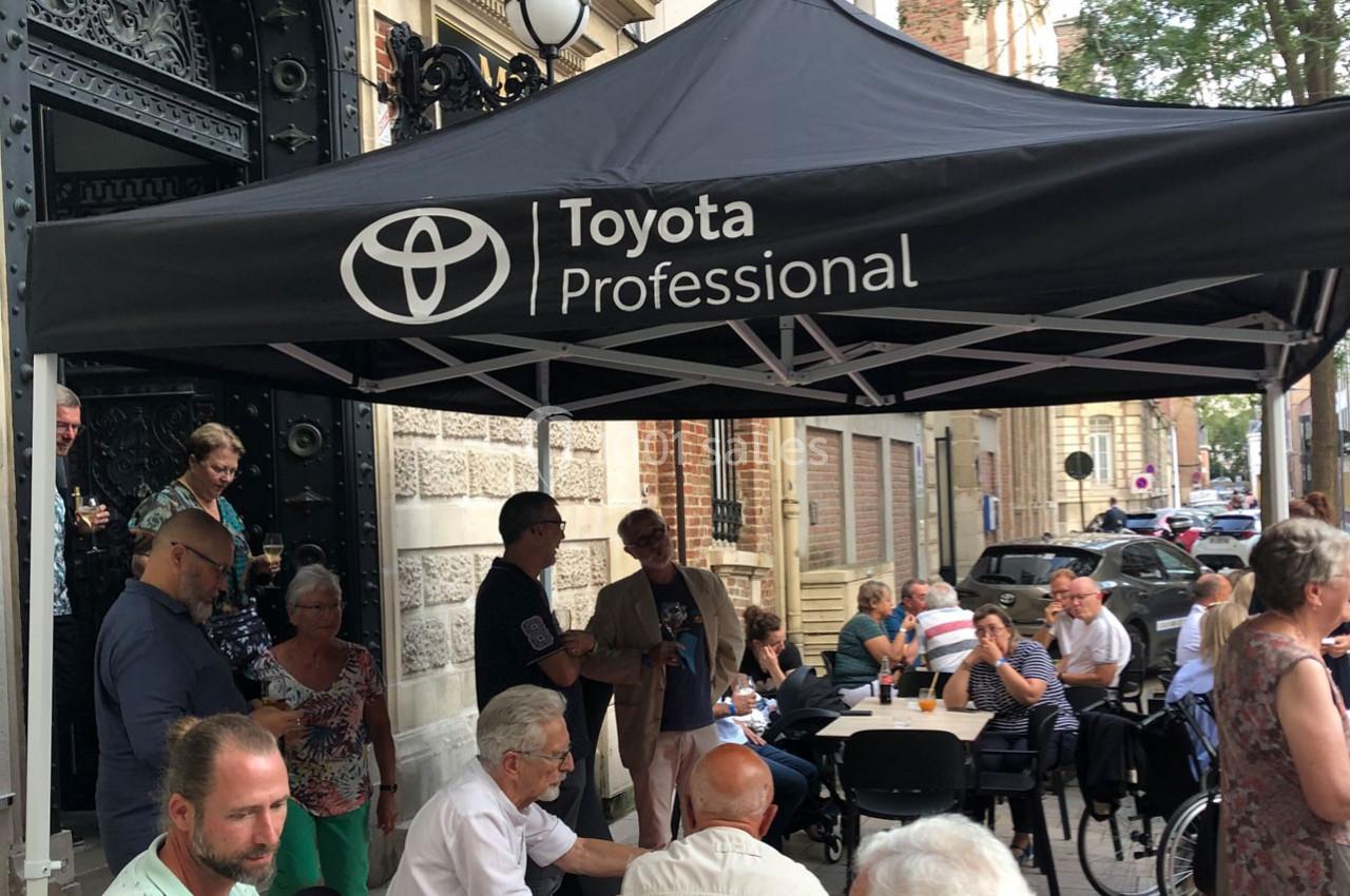 Groupe de personnes assises et debout sous un chapiteau noir marqué ’Toyota Professional’ dans une rue animée.