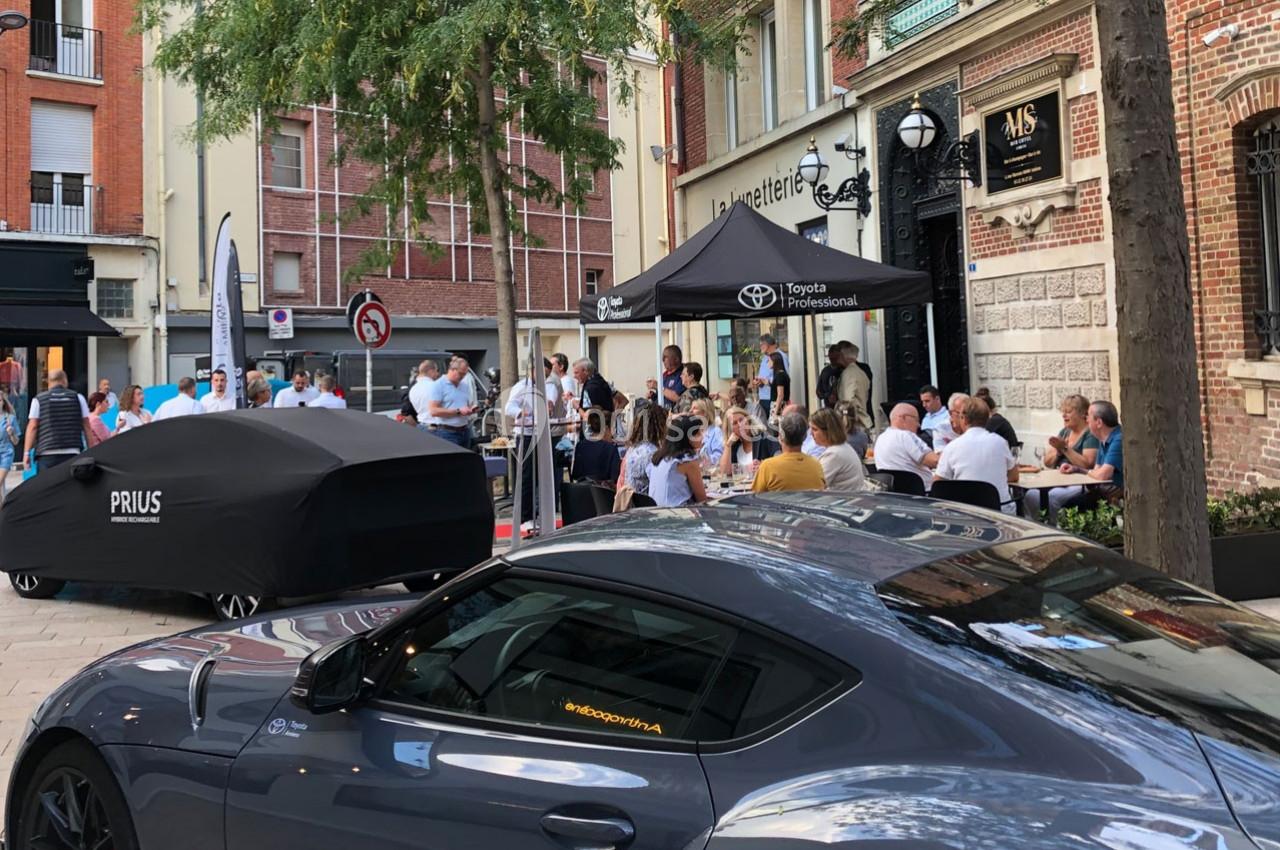 Voiture sportive grise stationnée devant un bâtiment, avec un événement rassemblant des personnes en arrière-plan.