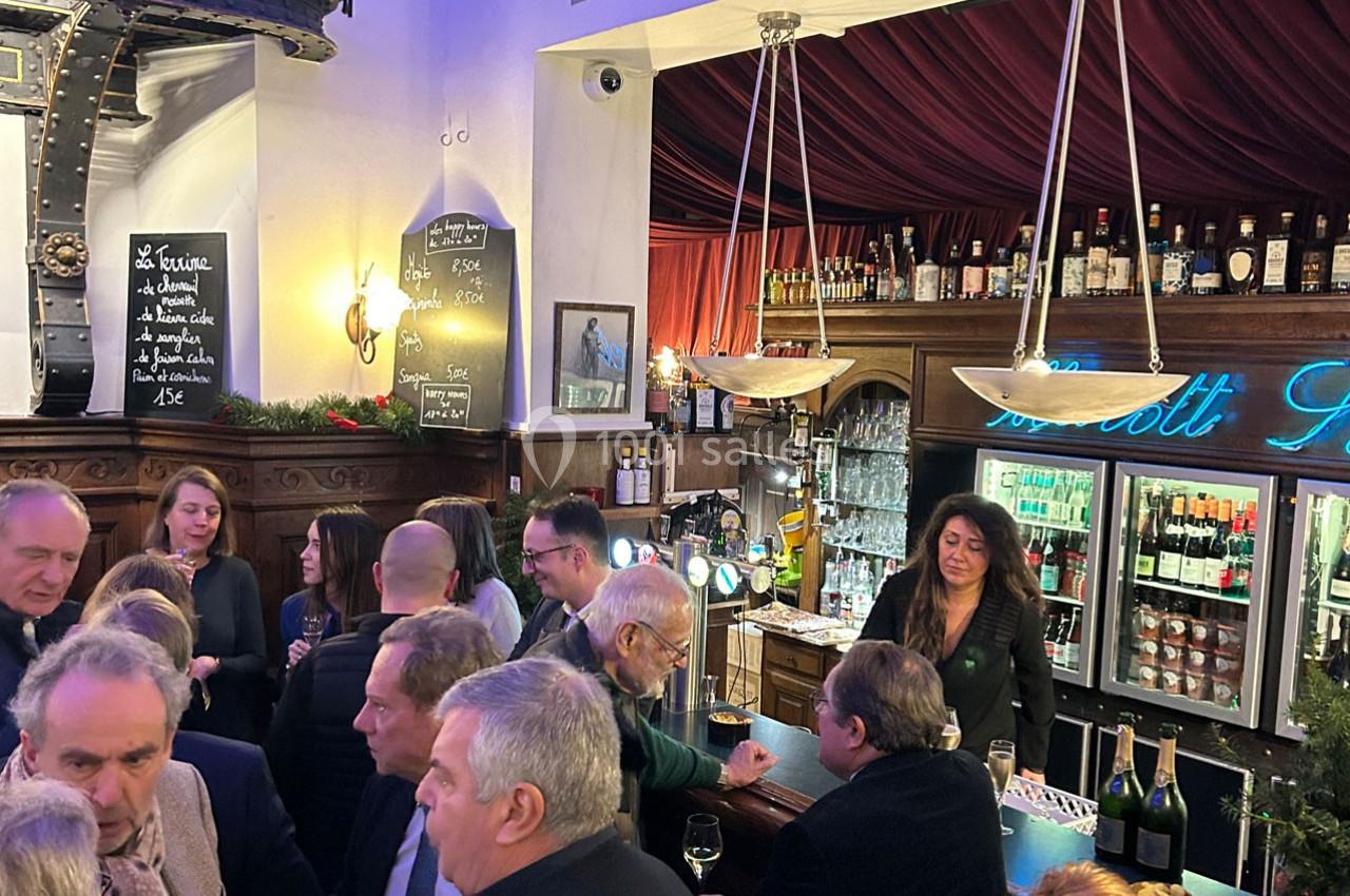 Groupe de personnes rassemblées dans un bar animé, avec une serveuse derrière le comptoir et des boissons exposées.