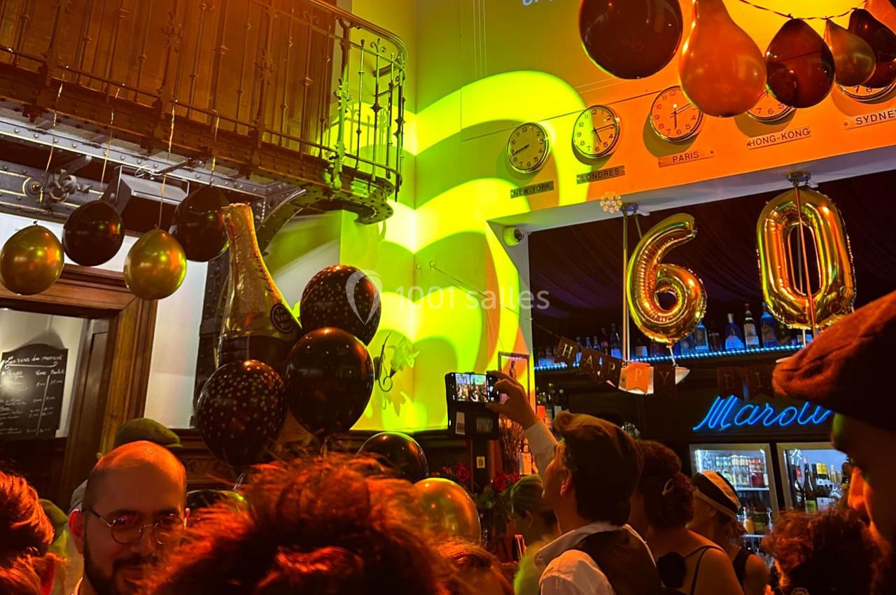 Foule célébrant un événement festif dans un bar décoré de ballons dorés et noirs, avec des horloges murales en arrière-plan.