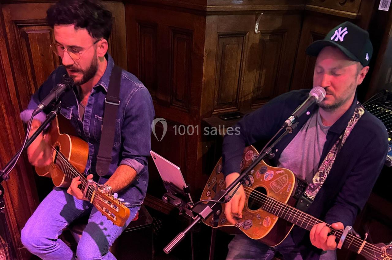 Deux musiciens jouent de la guitare et chantent dans un espace intérieur en bois, équipés de micros.