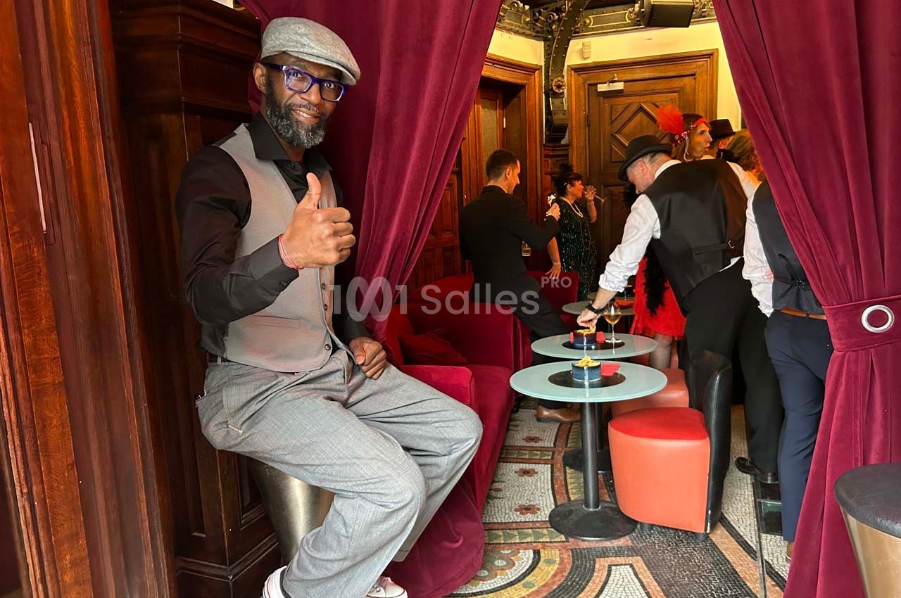 Un homme assis sur un tabouret, levant le pouce, dans un salon avec des rideaux rouges et des personnes en arrière-plan.
