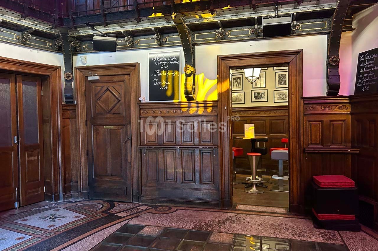Intérieur d'un bâtiment avec boiseries, carrelage au sol, et vue sur un espace avec tabourets rouges et tables.
