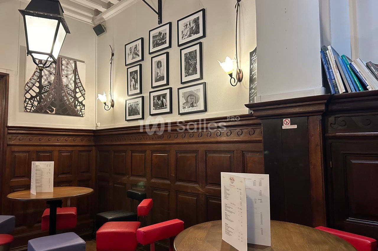 Intérieur d'un café avec tables, chaises colorées, murs en bois, photos encadrées et une grande lampe suspendue.