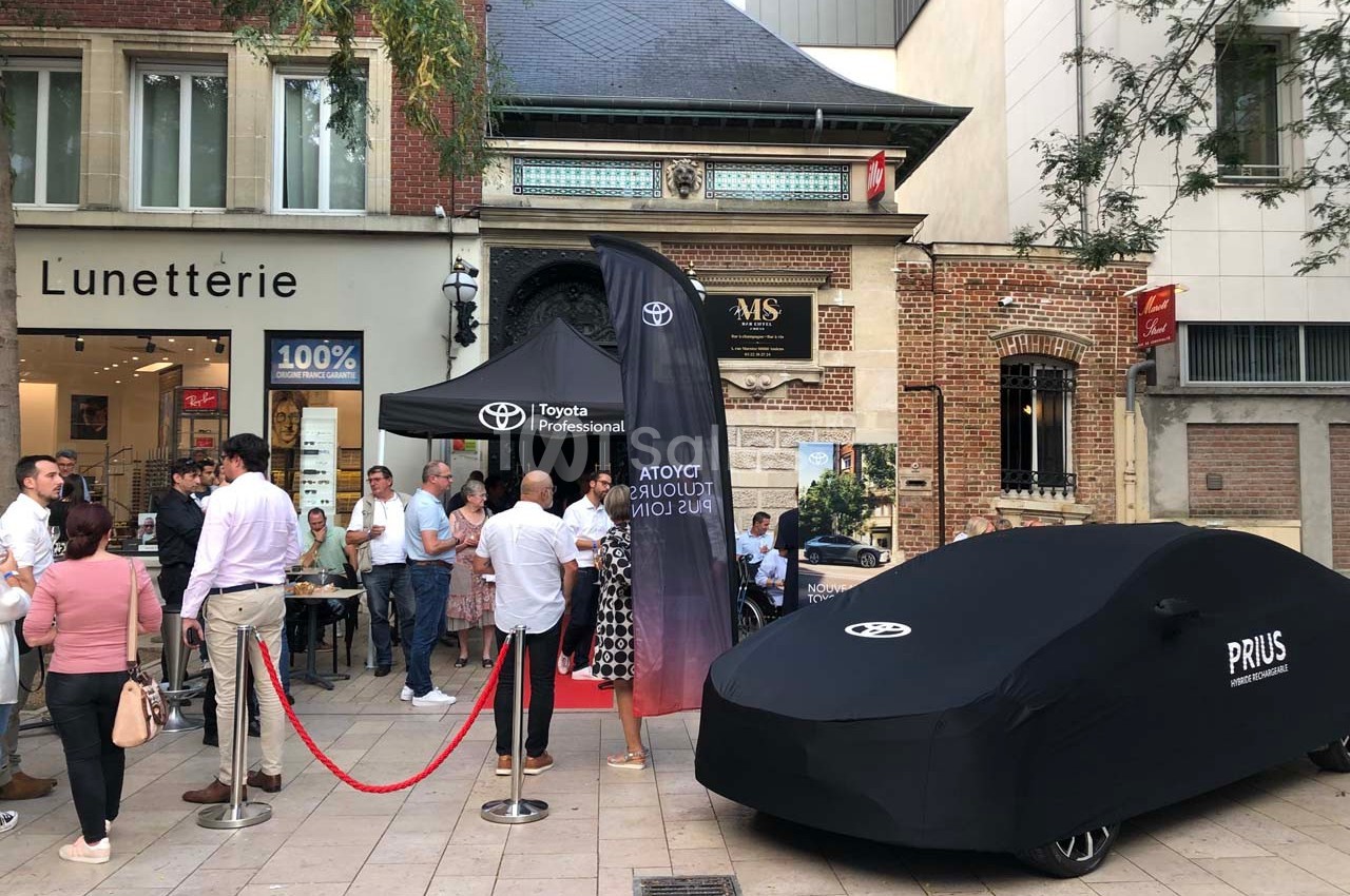Stand Toyota avec une voiture Prius couverte, situé sur une place devant des commerces et des passants.