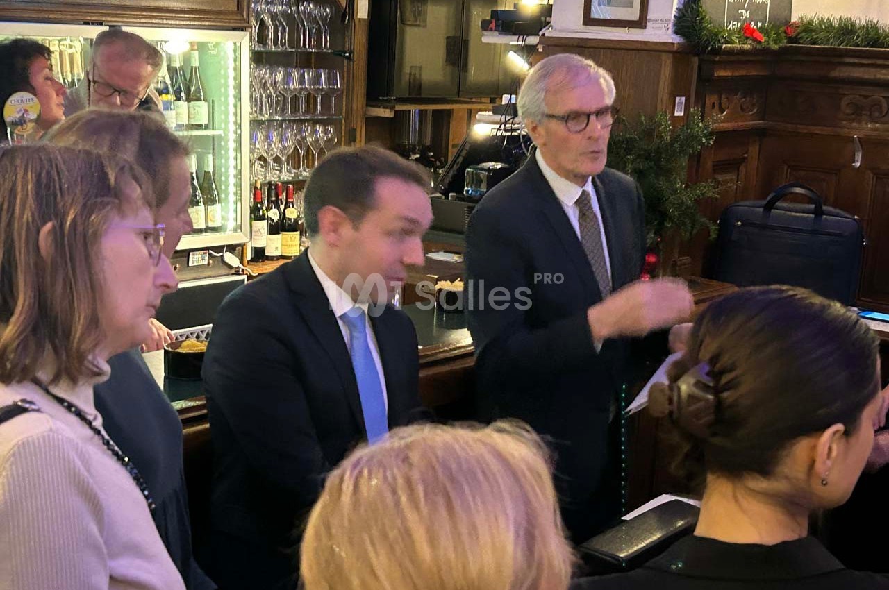 Des personnes discutent debout dans un bar ou restaurant, avec des bouteilles et des verres en arrière-plan.