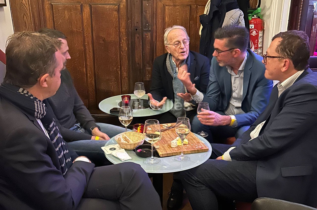 Cinq hommes discutent autour de petites tables avec des boissons et des collations dans un cadre intérieur chaleureux.