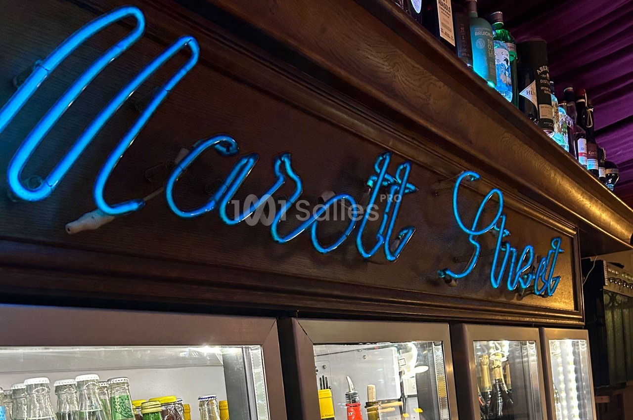 Enseigne lumineuse bleue ’Marott Street’ sur un meuble en bois, avec des bouteilles et un réfrigérateur en arrière-plan.