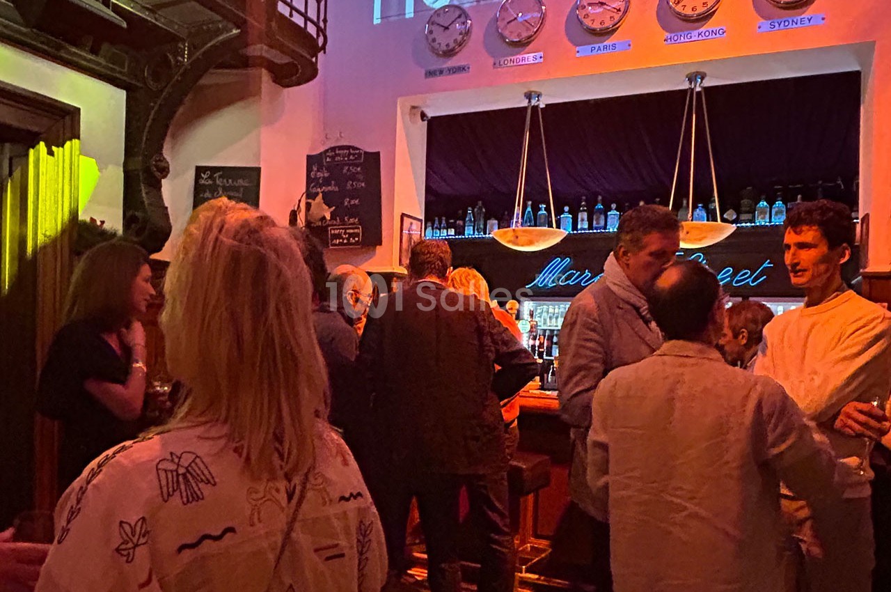 Des personnes discutent dans un bar à l'ambiance tamisée, avec une horloge murale affichant plusieurs fuseaux horaires.