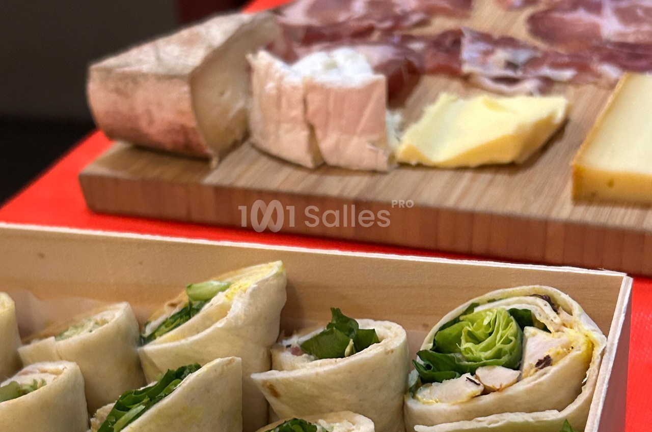 Plateau de wraps garnis de salade en premier plan, planche de charcuterie et fromages en arrière-plan.