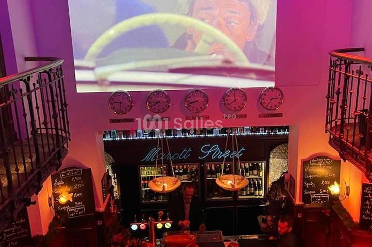 Vue intérieure d'un bar avec un écran projetant une scène de film, horloges murales et comptoir éclairé.