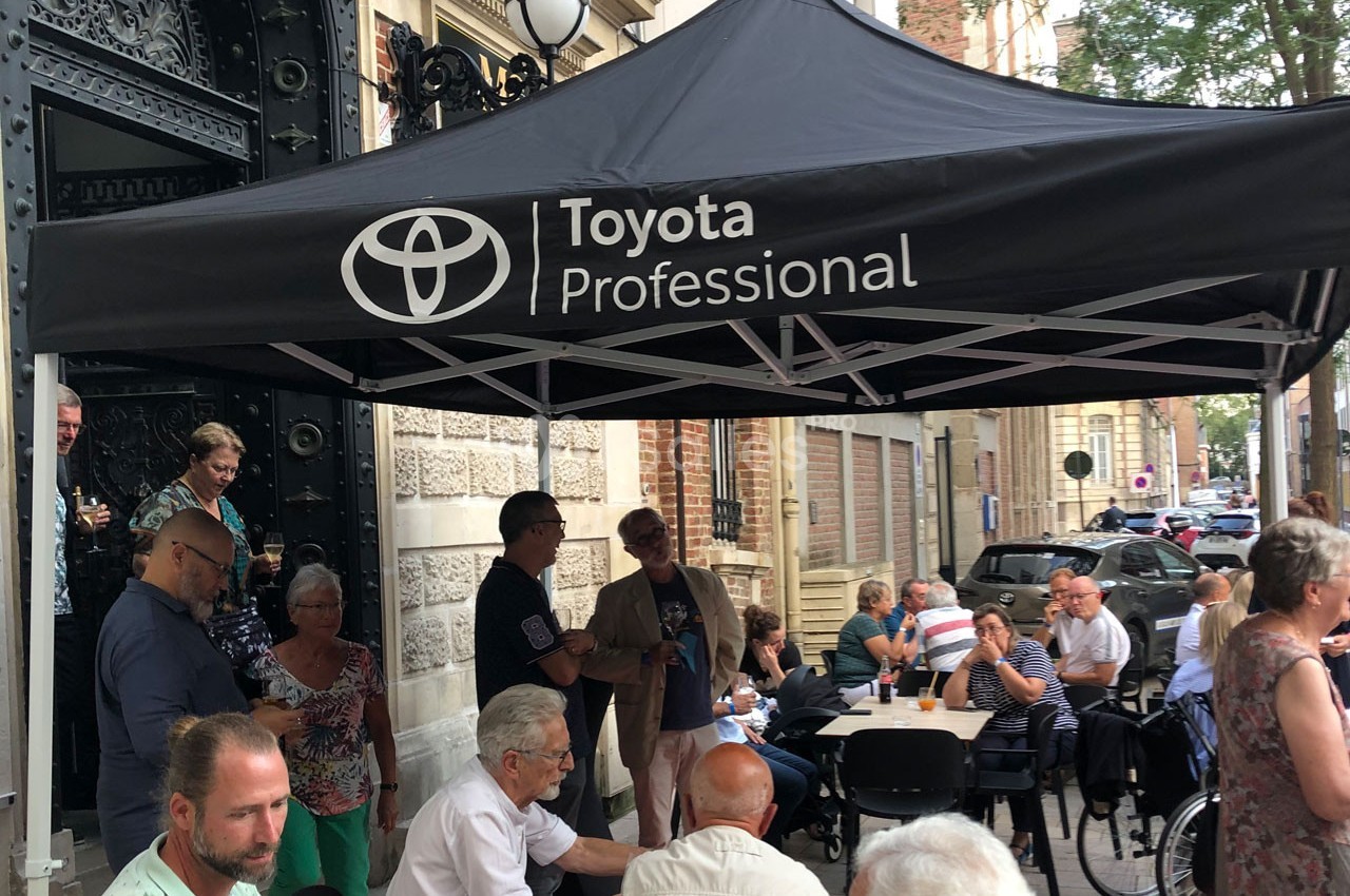 Un groupe de personnes assises et debout sous un auvent noir marqué ’Toyota Professional’ dans une rue animée.
