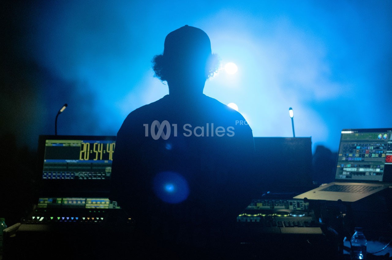 Un technicien de dos gère des équipements audio et lumineux dans une ambiance de scène éclairée en bleu.