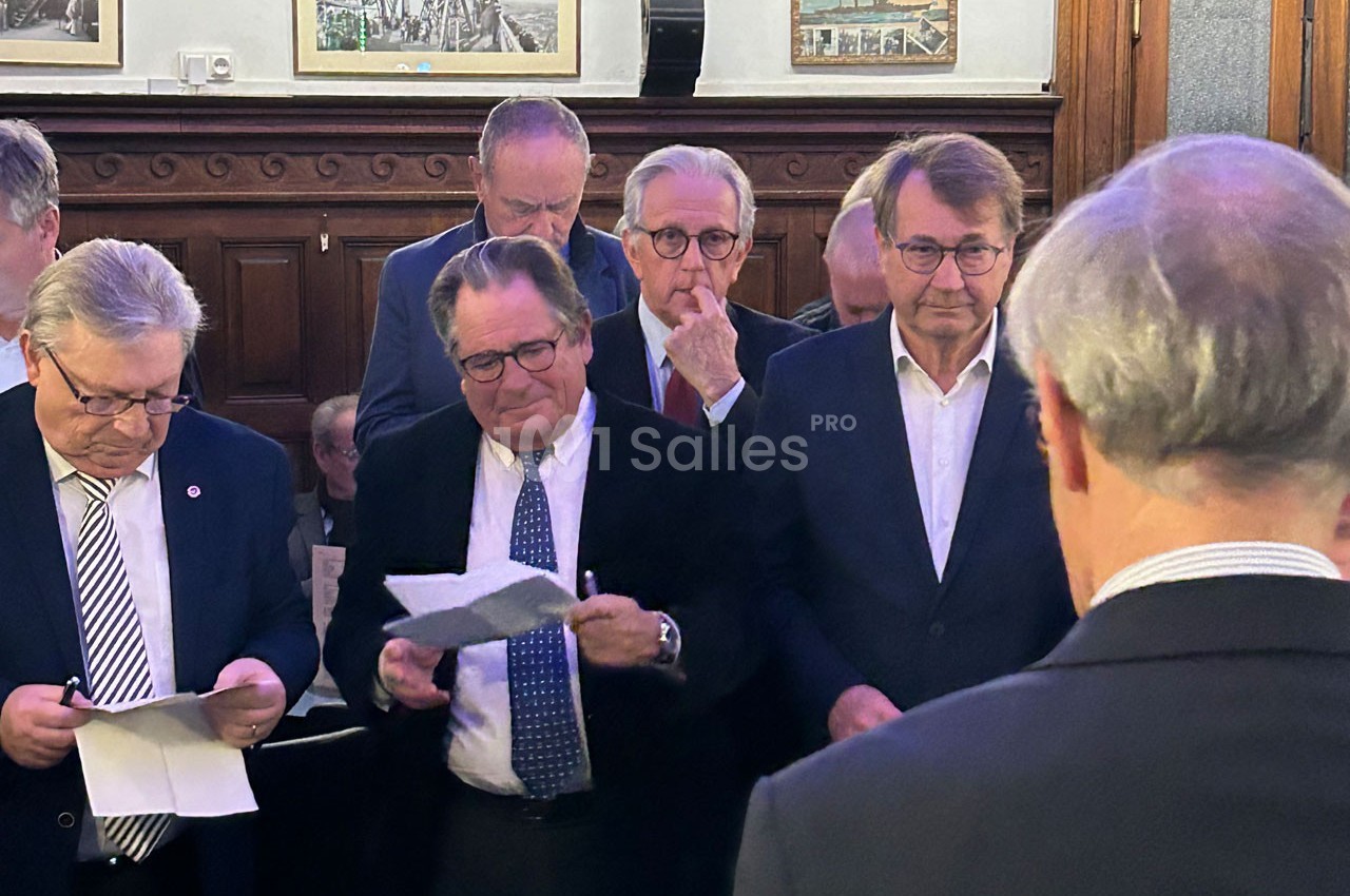 Des hommes en costume lisent des documents dans une salle avec des boiseries et des cadres accrochés au mur.