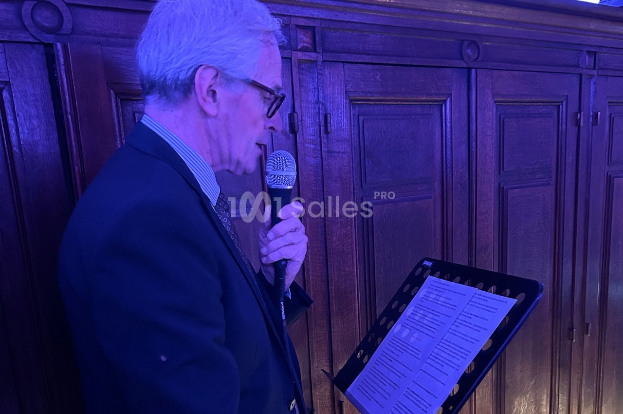Un homme âgé en costume lit un texte sur un pupitre tout en parlant dans un microphone, dans une pièce éclairée en bleu.