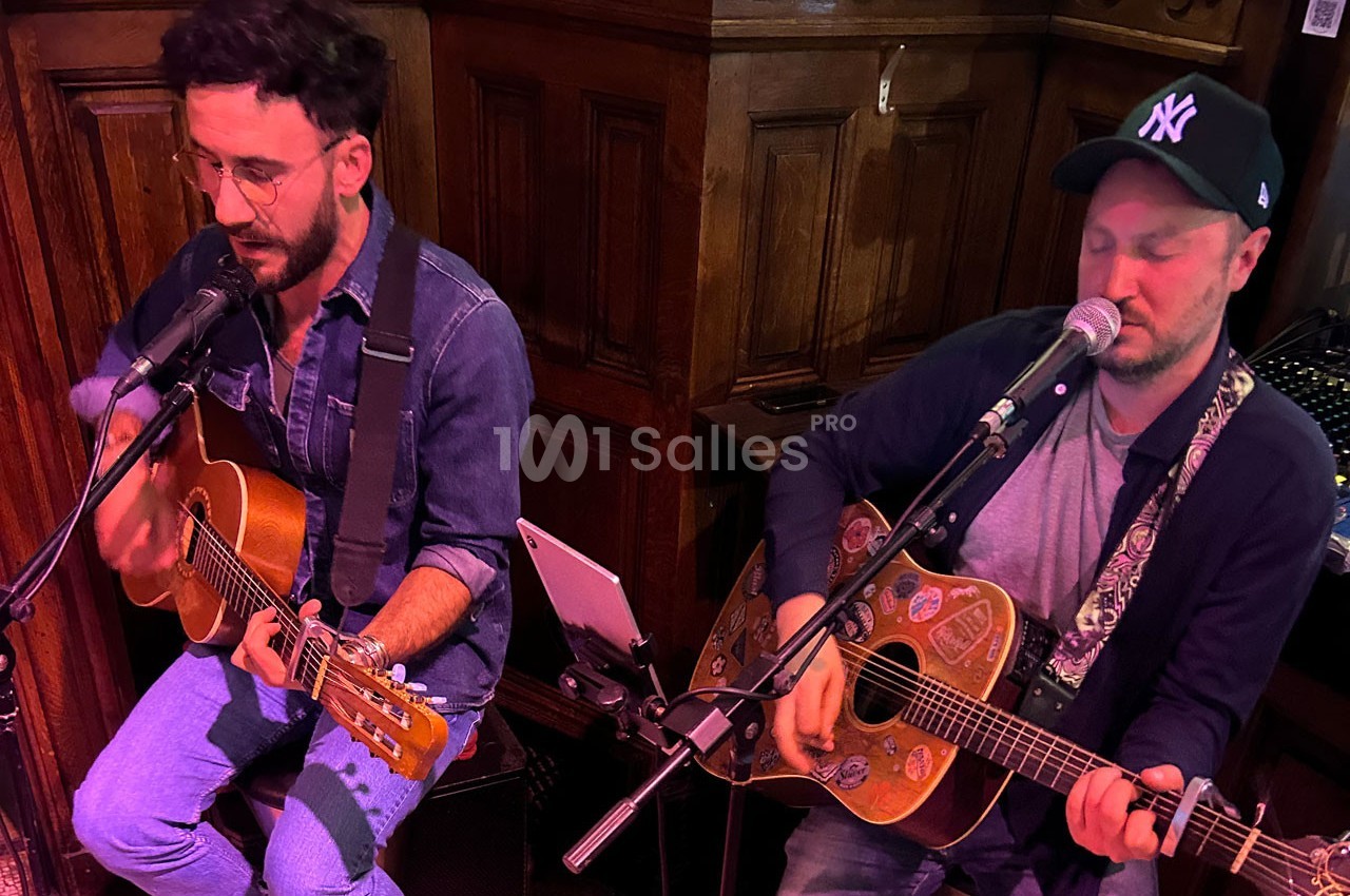 Deux musiciens jouent de la guitare et chantent dans un espace intérieur avec un décor en bois.