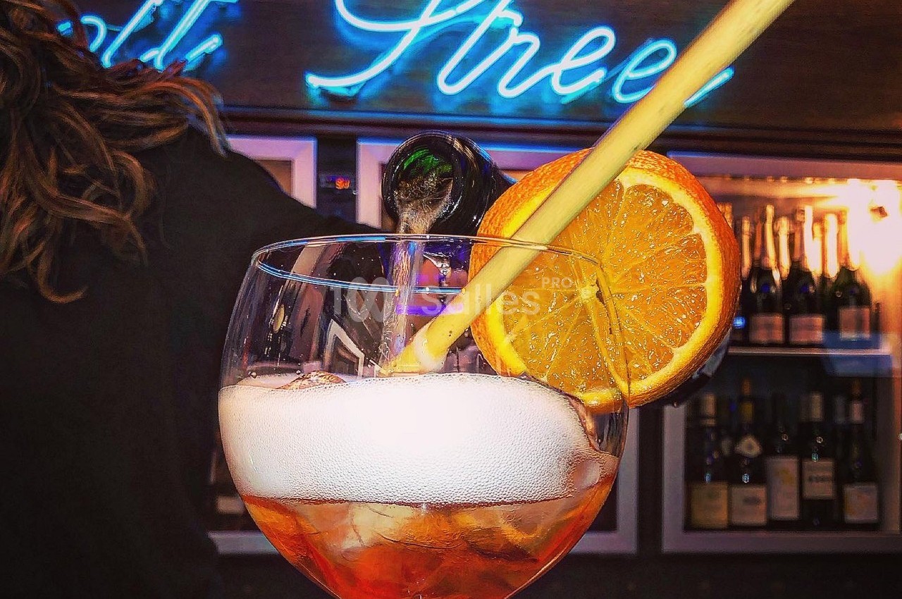 Verre de cocktail pétillant avec tranche d'orange et paille, servi devant un bar éclairé par un néon bleu.