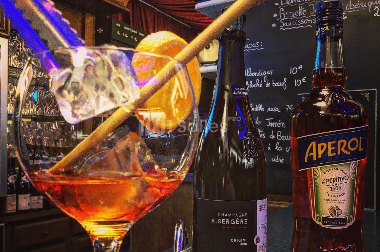 Verre de cocktail avec tranche d'orange, bouteille de champagne et Aperol sur un comptoir de bar.