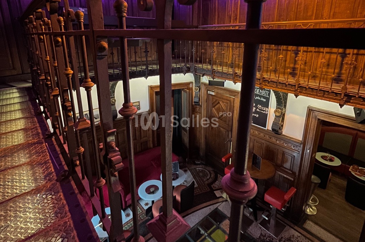 Vue intérieure d'un espace avec mezzanine, balustrade en métal, boiseries sculptées et tables basses éclairées.