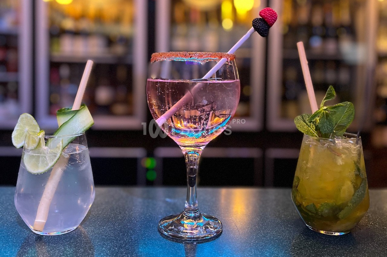 Trois cocktails colorés sur un bar, décorés de fruits, menthe et rondelles de citron, avec un fond flou de bouteilles.