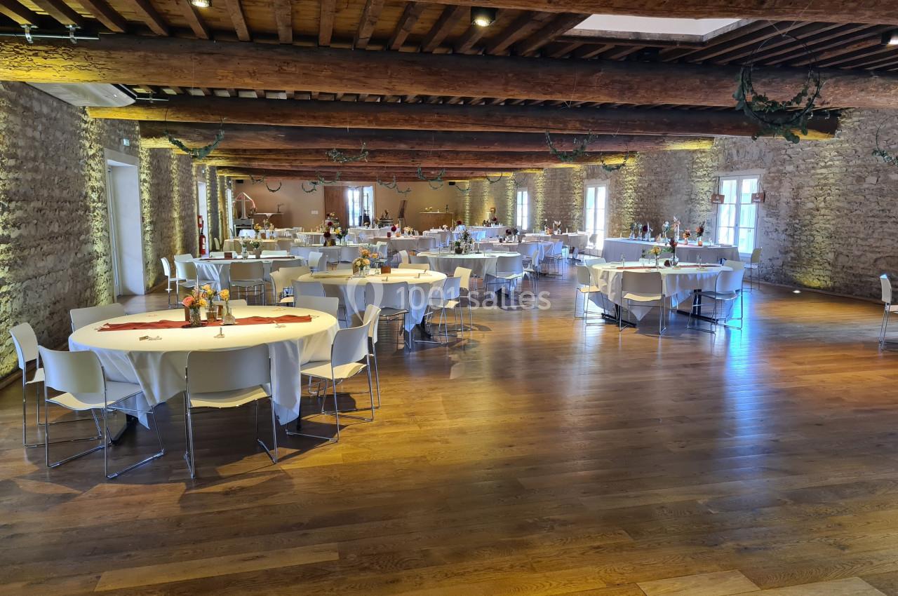 Salle de réception avec murs en pierre, plafond en poutres apparentes et tables rondes dressées pour un événement.