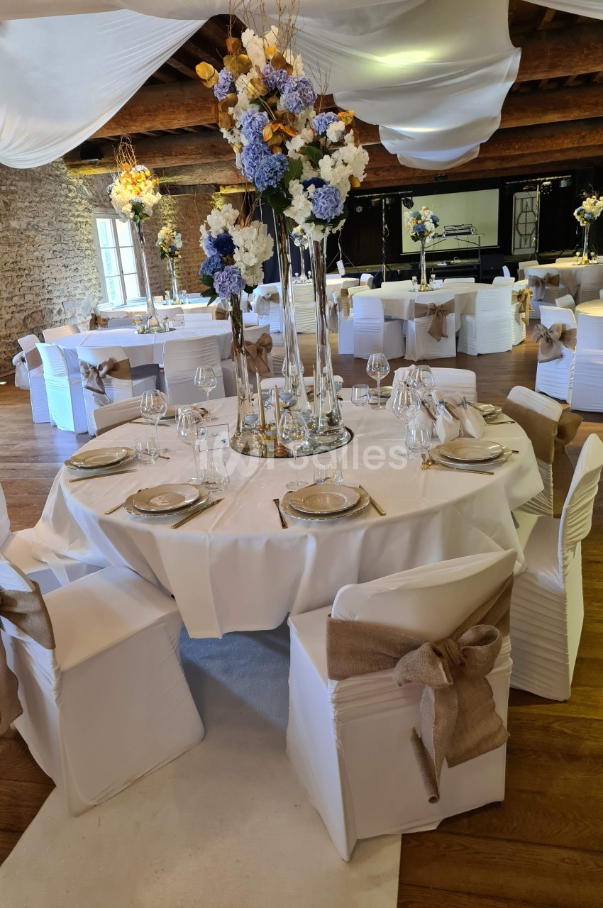 Salle de réception décorée avec des tables rondes, nappes blanches, chaises ornées de nœuds en toile de jute et centres de…