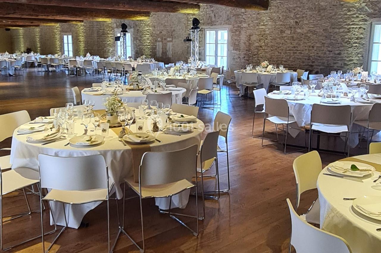 Salle de réception avec des tables rondes dressées, nappes blanches, chaises modernes et murs en pierre apparente.