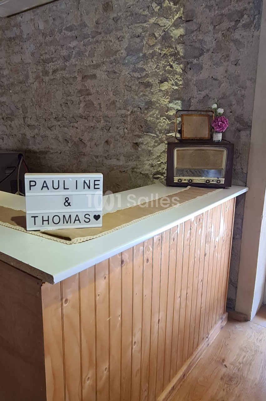 Comptoir en bois avec une boîte lumineuse affichant ’Pauline & Thomas’, une radio vintage et un vase de fleurs.