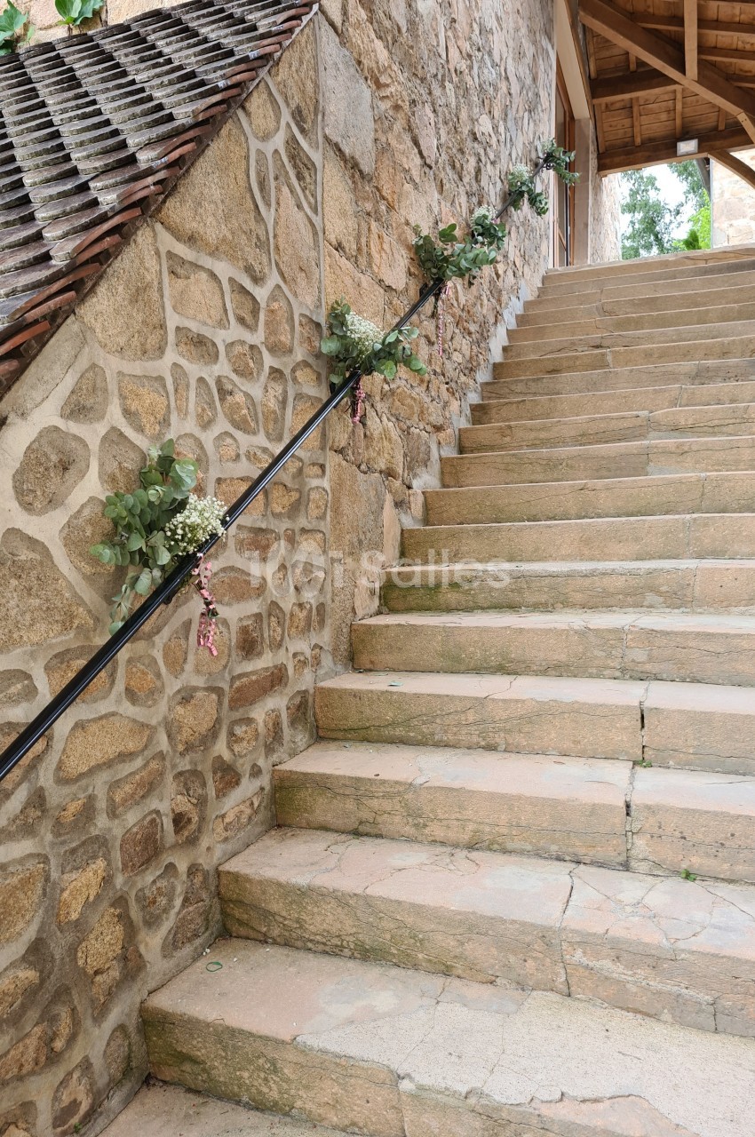 Escalier en pierre bordé d'un mur en pierre décoré de fleurs et verdure, menant à une entrée sous un toit en bois.