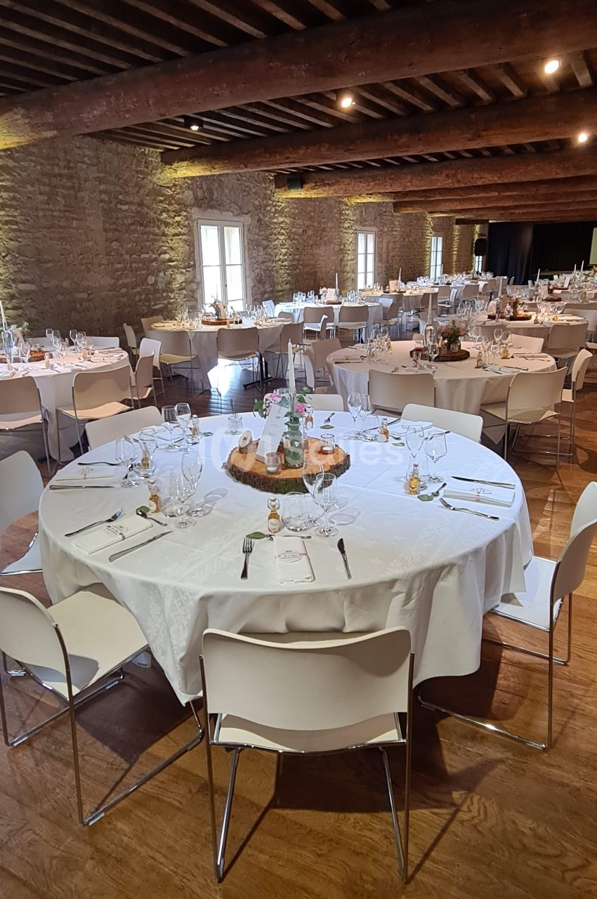 Salle de réception avec tables rondes dressées, nappes blanches, chaises modernes et murs en pierre apparente.