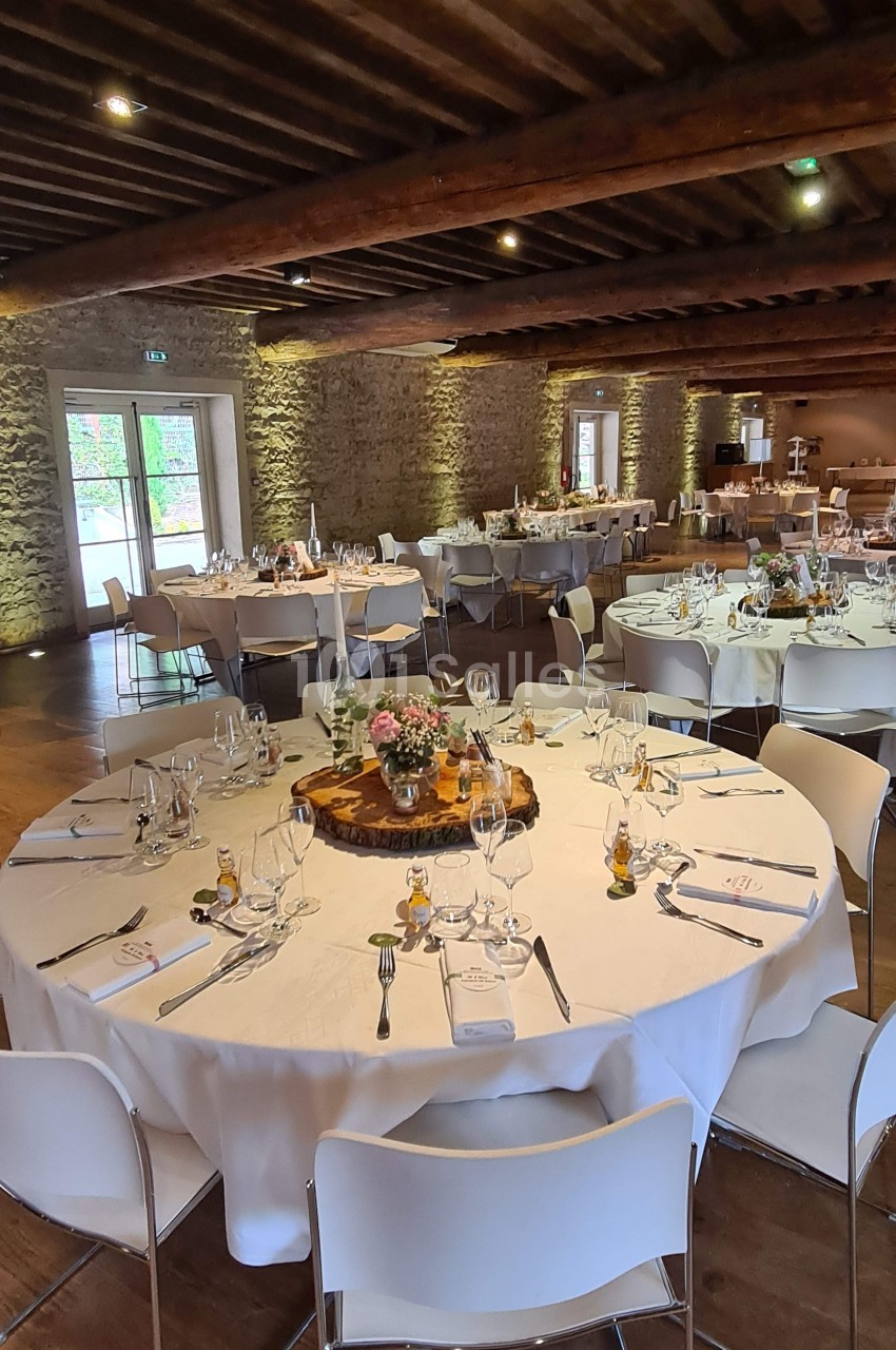 Salle de réception avec tables rondes dressées, nappes blanches, décorations florales et murs en pierre apparente.