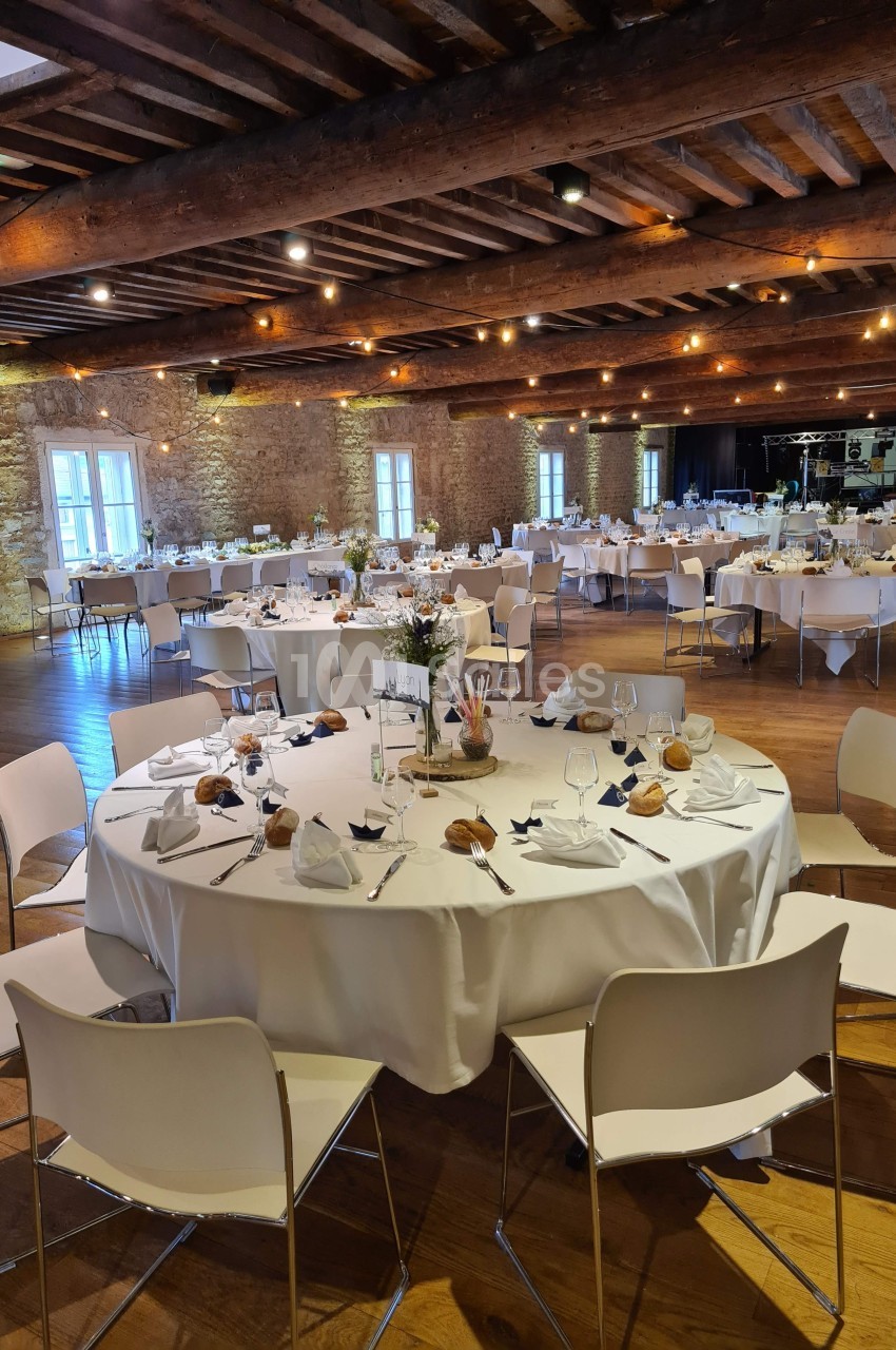 Salle de réception avec tables rondes dressées, nappes blanches, chaises modernes et éclairage chaleureux.