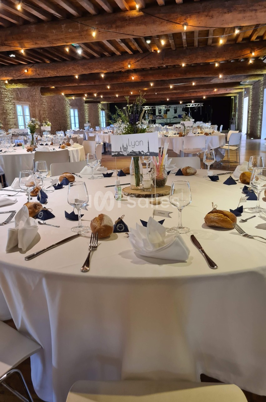 Salle de réception avec tables rondes dressées, nappes blanches, couverts, verres, pains et décorations florales.