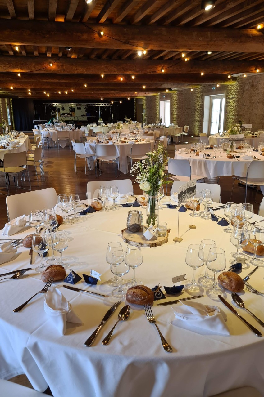 Salle de réception décorée avec des tables rondes dressées, nappes blanches, vaisselle élégante et bouquets de fleurs.