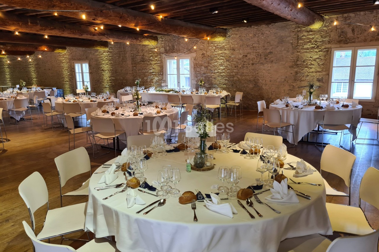 Salle de réception en pierre avec tables rondes dressées pour un repas, décorée de fleurs et éclairée par des guirlandes…