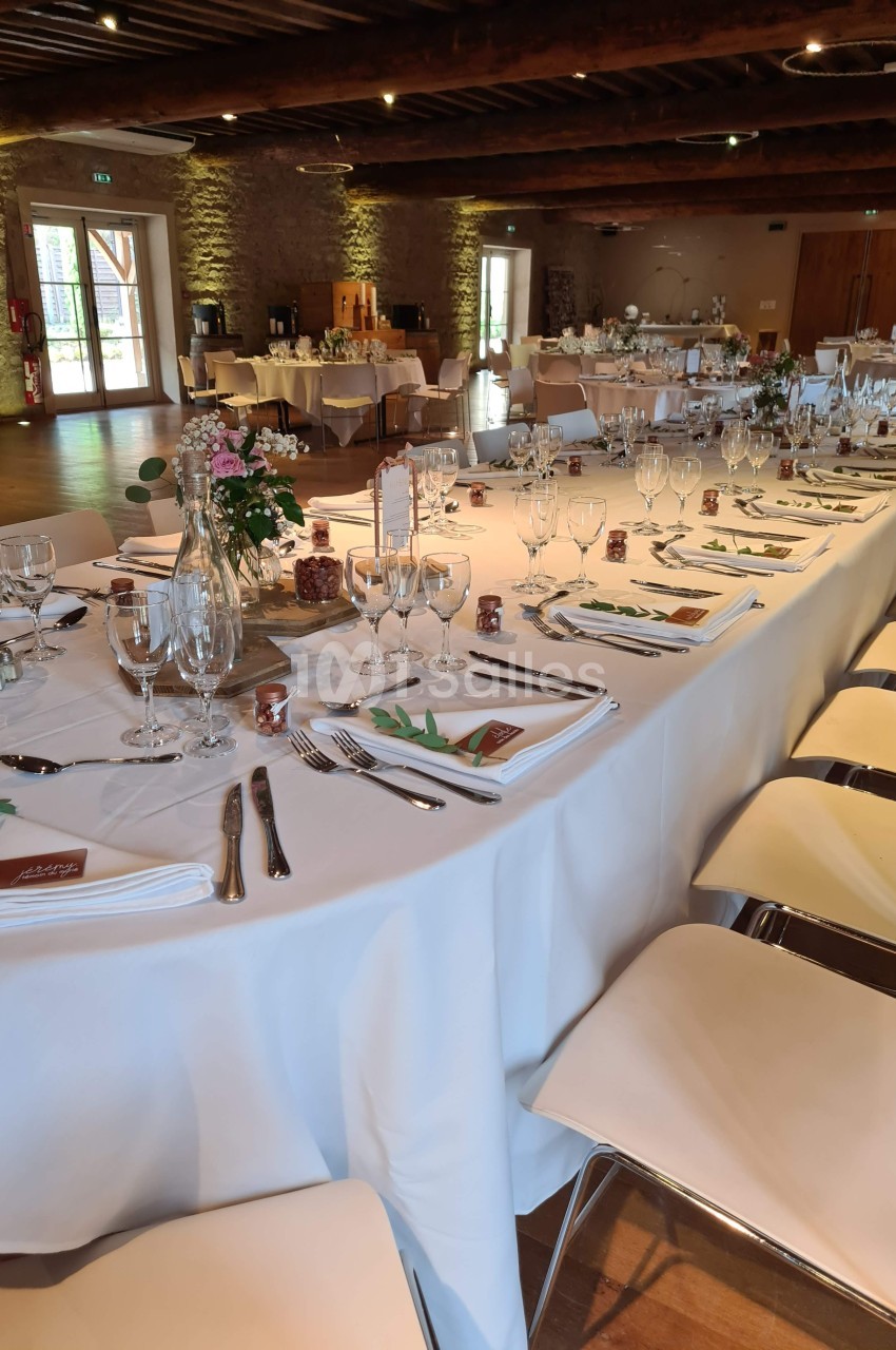 Salle de réception avec longues tables dressées, nappes blanches, vaisselle élégante et décoration florale.