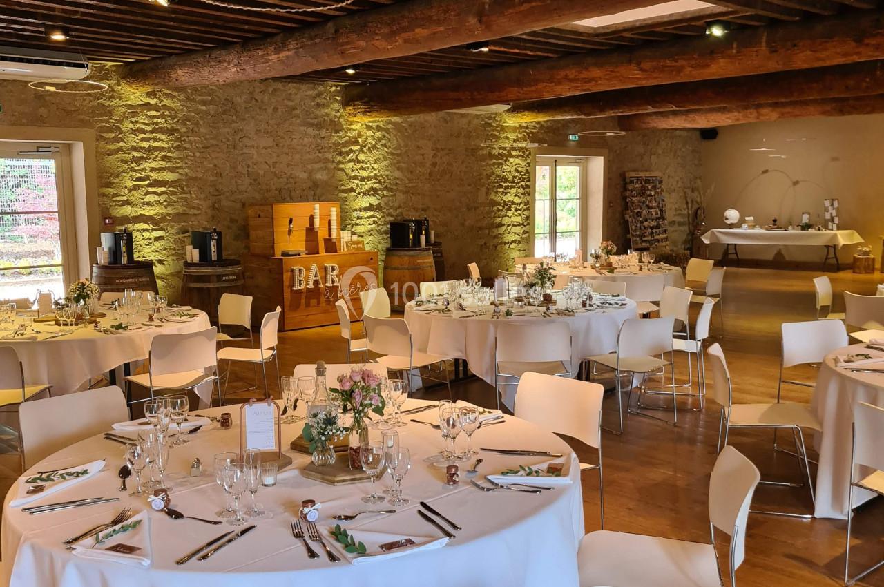 Salle de réception avec tables rondes dressées, chaises blanches et décor rustique en bois et pierre apparente.