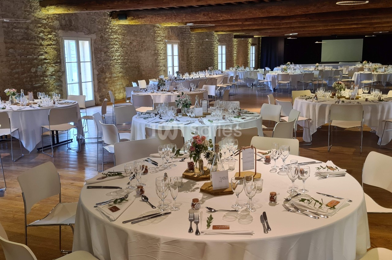 Salle de réception décorée avec des tables rondes dressées pour un événement, éclairée par la lumière naturelle.
