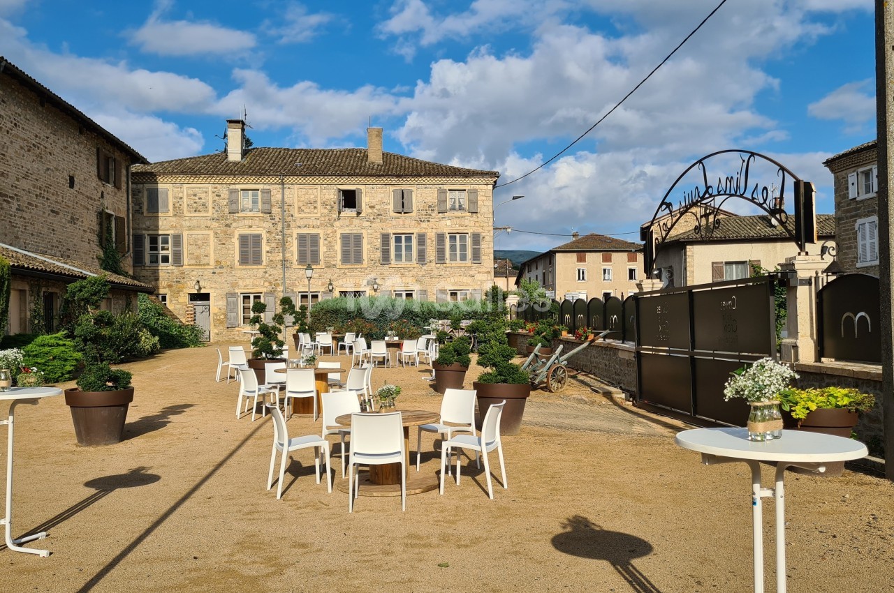 Cour extérieure avec tables et chaises blanches, entourée de bâtiments en pierre sous un ciel partiellement nuageux.