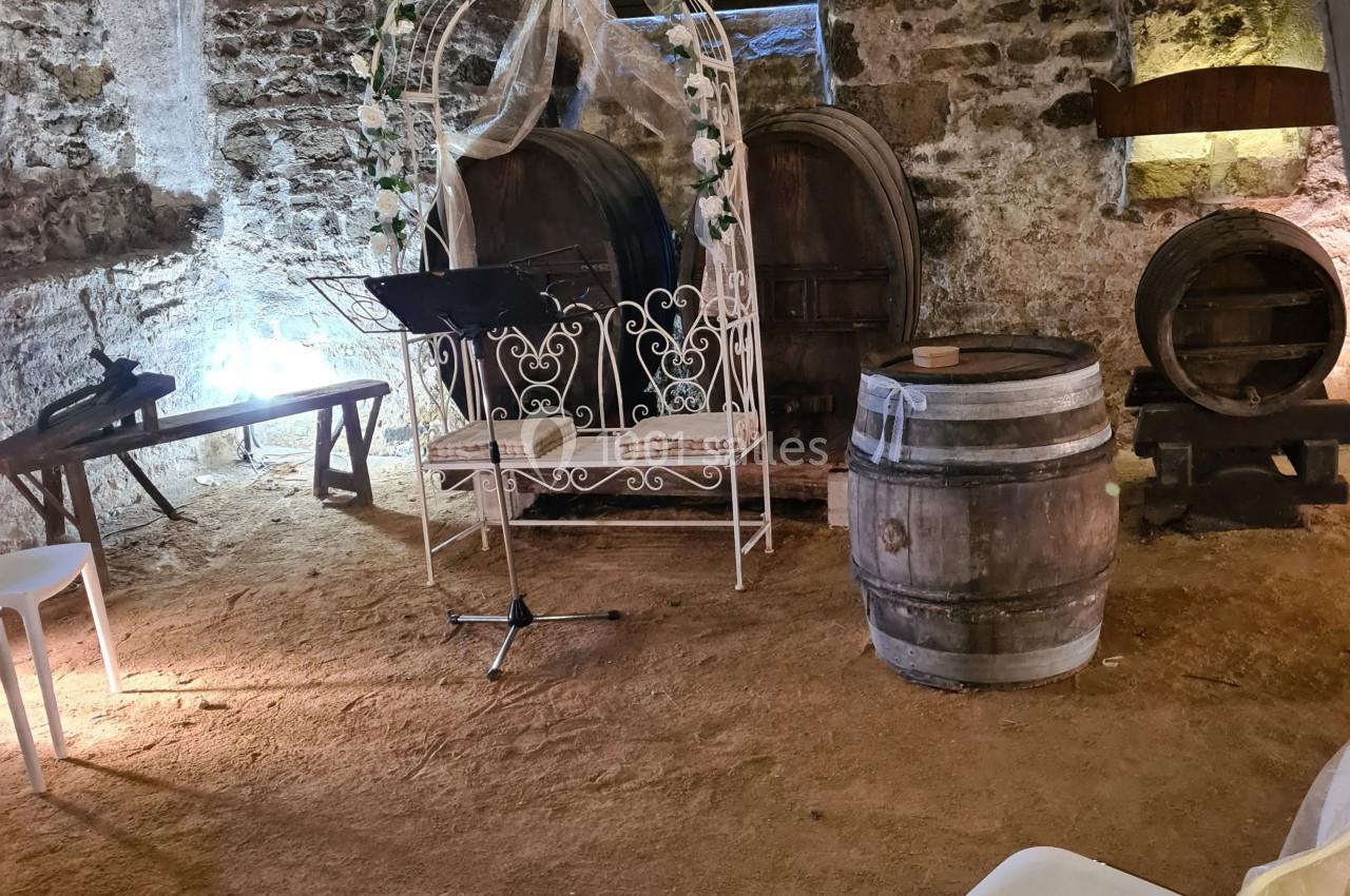 Salle voûtée en pierre avec tonneaux, arche décorée de fleurs, pupitre et chaises blanches disposées au sol.