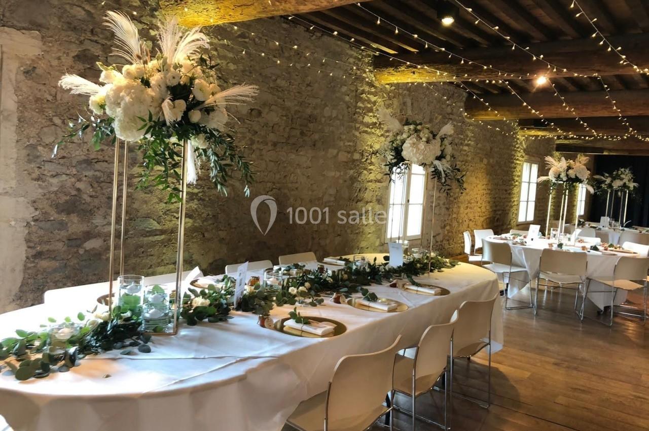 Salle de réception décorée pour un mariage, avec tables blanches ornées de fleurs et guirlandes lumineuses suspendues.