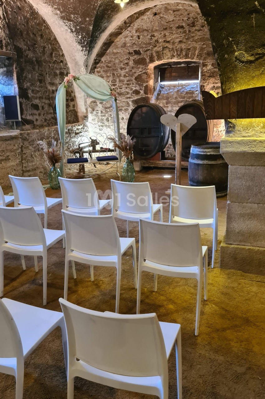 Salle voûtée en pierre aménagée pour une cérémonie, avec chaises blanches alignées et une arche décorative au fond.